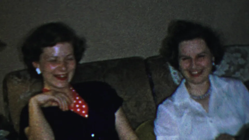 Two adult women laugh together on a living-room couch in the supplied 1954 Columbus, Ohio home-movie footage, with one woman in a dark top and red polka-dot collar and another in a white blouse.