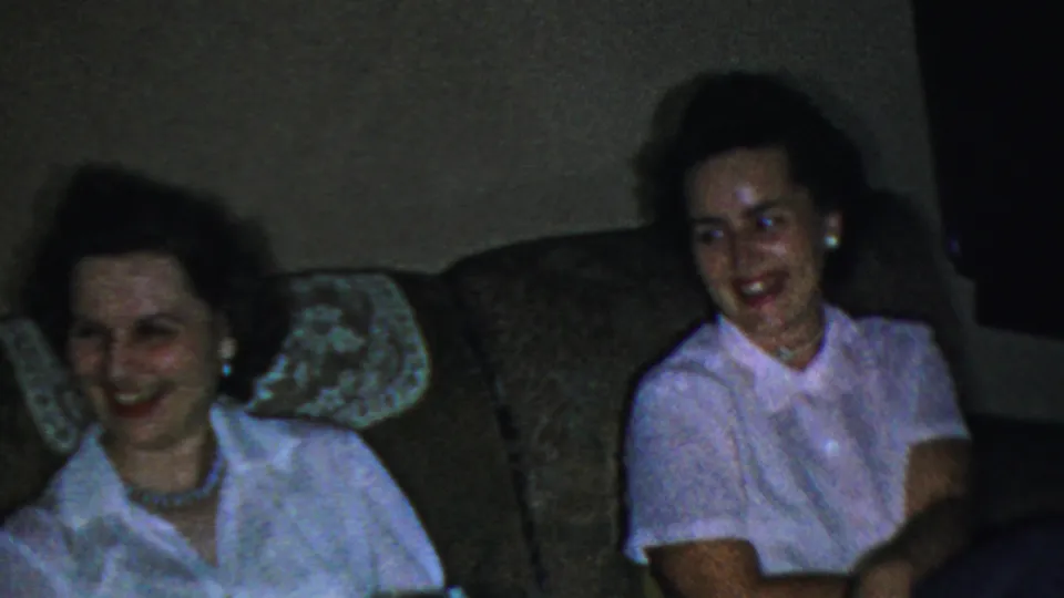 The 1954 Columbus, Ohio home-movie frame shows two women seated together on a couch, one in a white blouse and one partly shadowed, while another woman in a light pink shirt appears at the right edge.