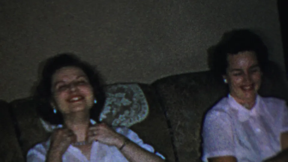 Adult woman in a white collared shirt and pearl necklace laughs on an upholstered couch in a Columbus, Ohio home-movie interior from the 1954 clip.