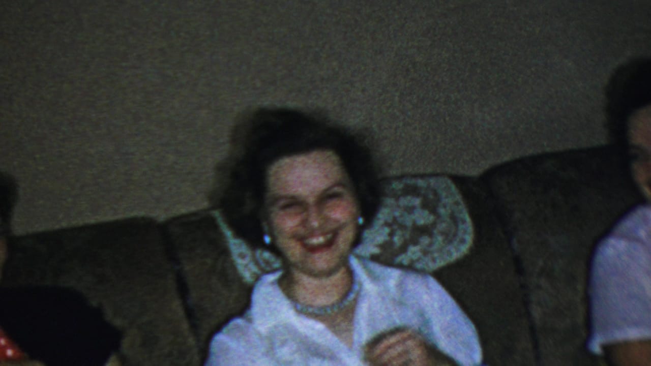 Adult woman in a white collared shirt and pearl necklace smiles broadly while seated on a couch in the 1954 Columbus, Ohio home-movie living-room scene.
