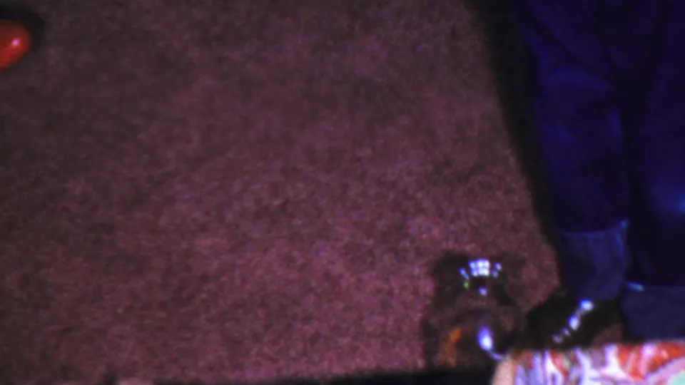 1954 Columbus, Ohio home-movie frame showing the game bottle on brown carpet near a person's dark pants and black shoes, with the subject mostly cropped out.
