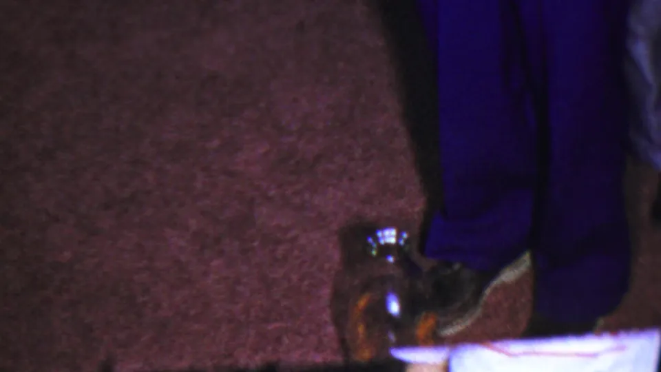 In the Columbus, Ohio 1954 home-movie frame, a reflective glass object sits near a dark shoe on burgundy carpet while a pale cloth or paper crosses the bottom edge.
