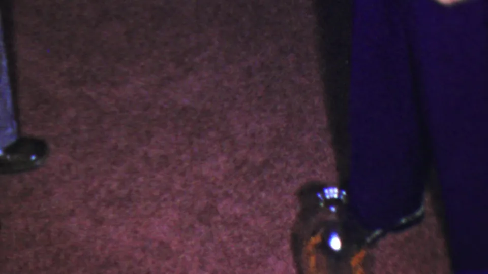 Cropped 8mm frame showing a brown glass bottle on a dark carpet beside partially visible children during the indoor dropping game.