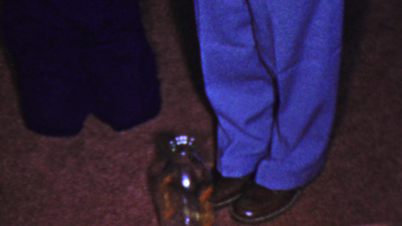 1954 Columbus, Ohio home-movie frame showing two people cropped at the legs, with bright blue trousers, dark pants, brown shoes, and a reflective glass jar on the carpet.