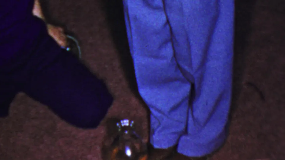 Home-movie frame showing a dark-clothed person low to the carpet near a reflective glass jar, with another person standing in bright blue trousers beside them.