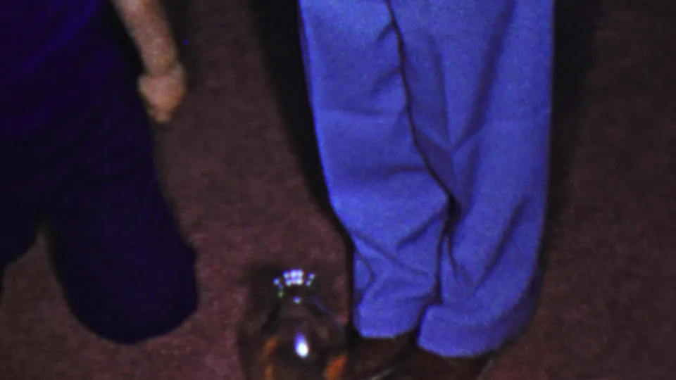 Final frame of the brief 1954 Columbus home-movie moment, with a dark-clothed figure low on the carpet, standing blue-trousered legs, and a glass jar reflecting light.