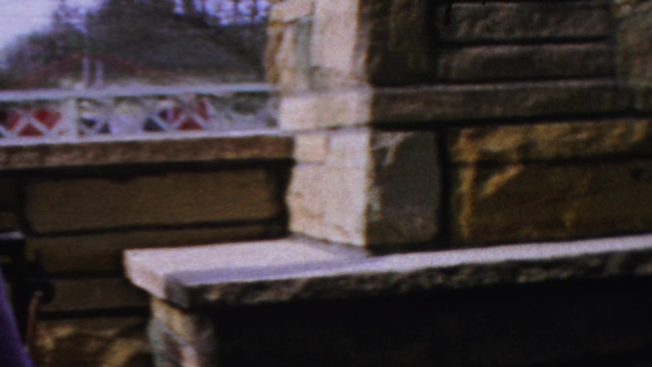 A 1954 Columbus, Ohio 8mm frame shows a stone outdoor fireplace area with a patterned railing, bench, and partial figure at the edge.