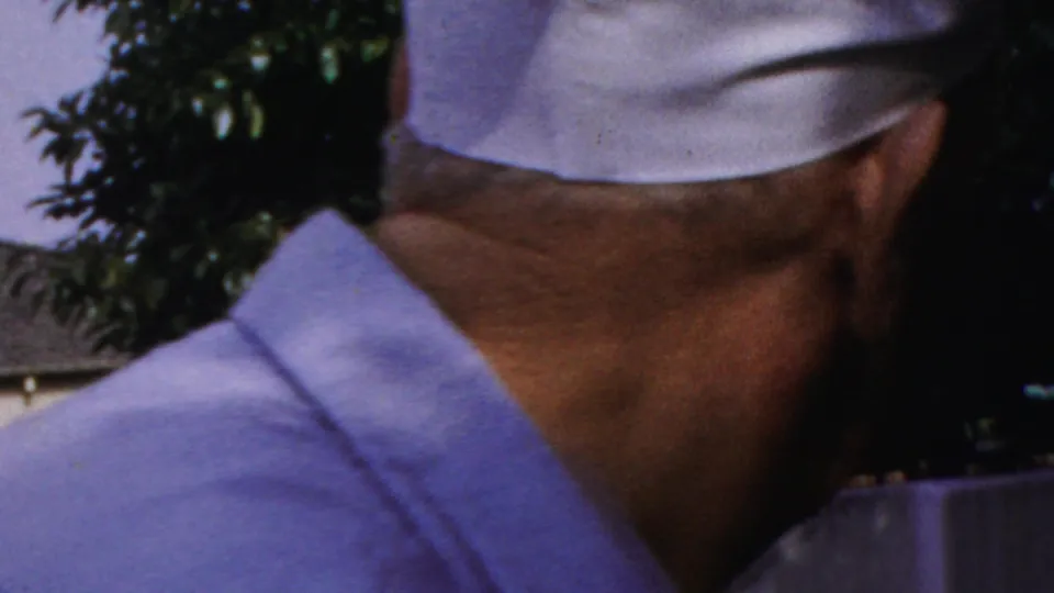 In a 1954 Columbus, Ohio home-movie frame, two older men in white caps stand outdoors near trees and a wall, with one man partly turned away and the other facing the camera.