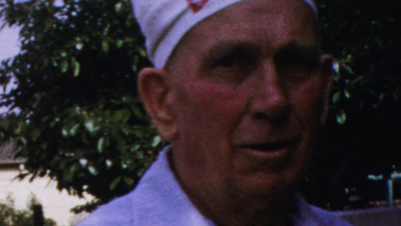 Older man in a white cap and light shirt faces the camera near leafy trees.