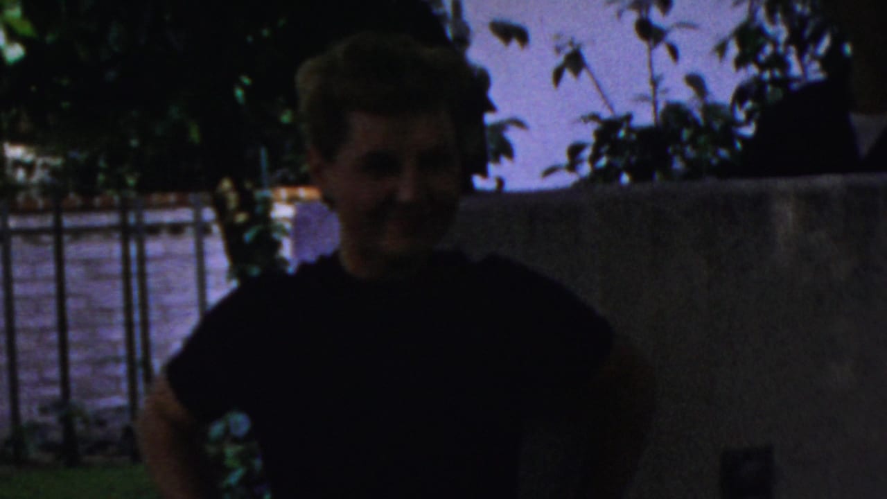 In 1954 Columbus home-movie footage, a blonde woman in a dark top stands in shadow near leafy branches and a low wall, with a bright background beyond.