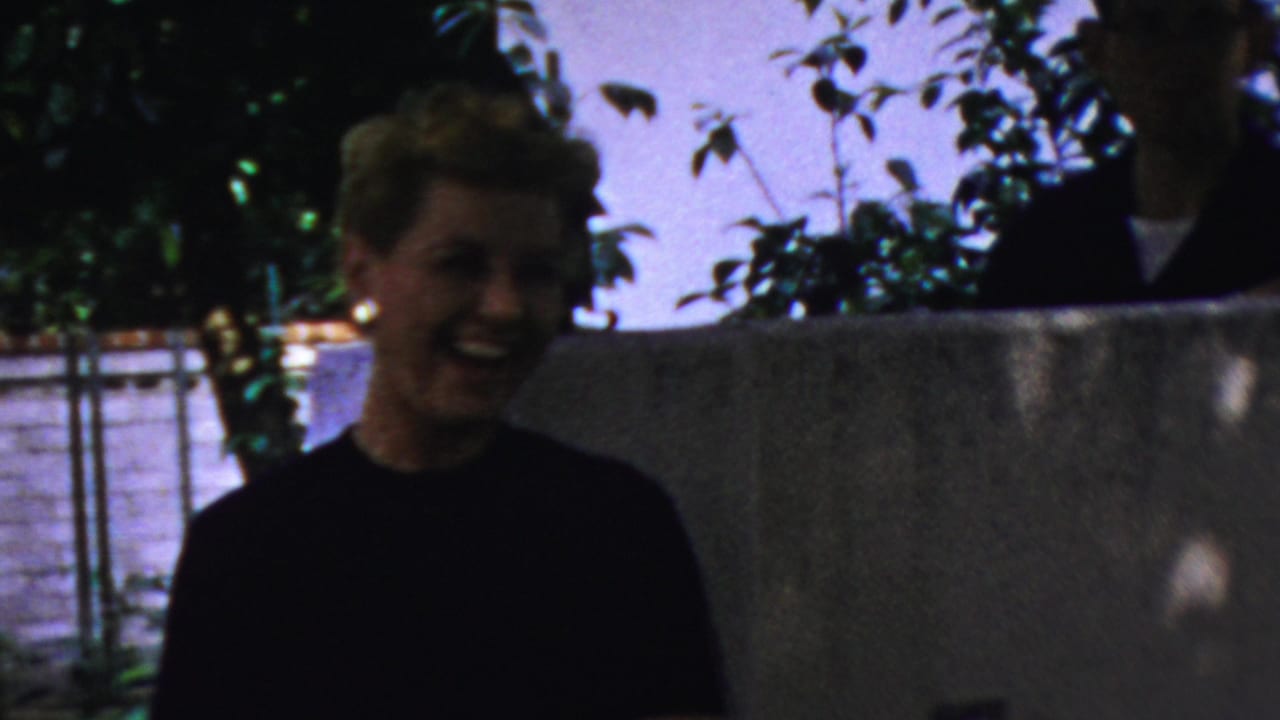 Smiling blonde woman outdoors with foliage and a low wall behind her.