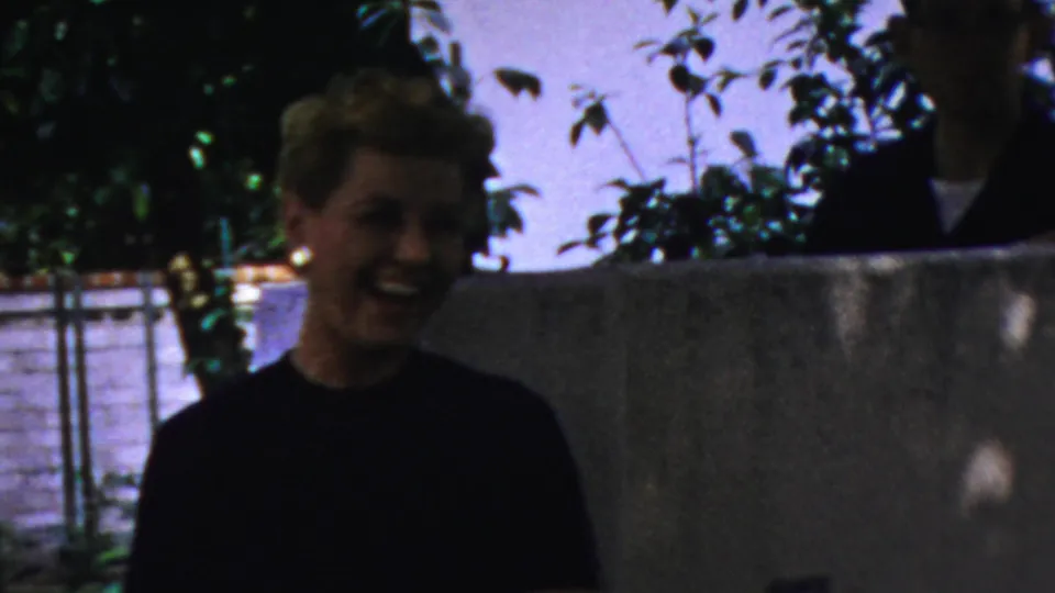 A smiling blonde woman in a dark top appears outdoors in the supplied Columbus, Ohio footage, framed beside foliage and a low wall under uneven natural light.