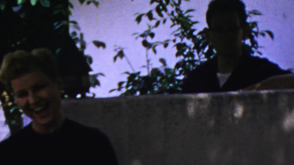 A laughing blonde woman is framed low in the foreground while a shadowed adult man stands behind the wall in this 1954 Columbus 8mm home-movie frame.