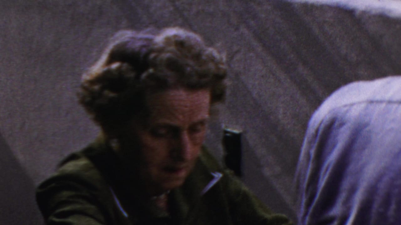 Curly-haired woman in a green jacket looks downward near a person in a light blue shirt.