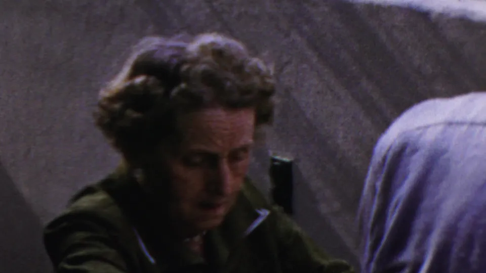 The woman appears in closer view during the 1954 Columbus family gathering, looking down or speaking near a foreground adult in a light blue shirt.