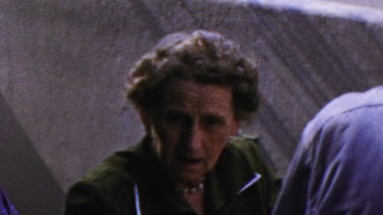 A close handheld frame shows the woman facing toward the camera area, with another adult in a light blue shirt partly obscuring the right side of the gathering.