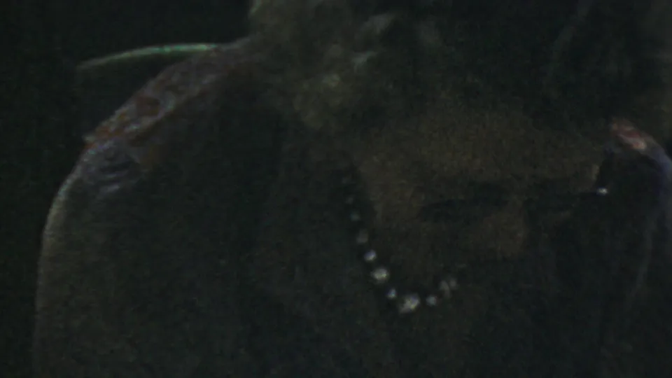Older woman wearing glasses and a pearl necklace looks downward in a dark, soft 1954 Columbus, Ohio home-movie close-up.
