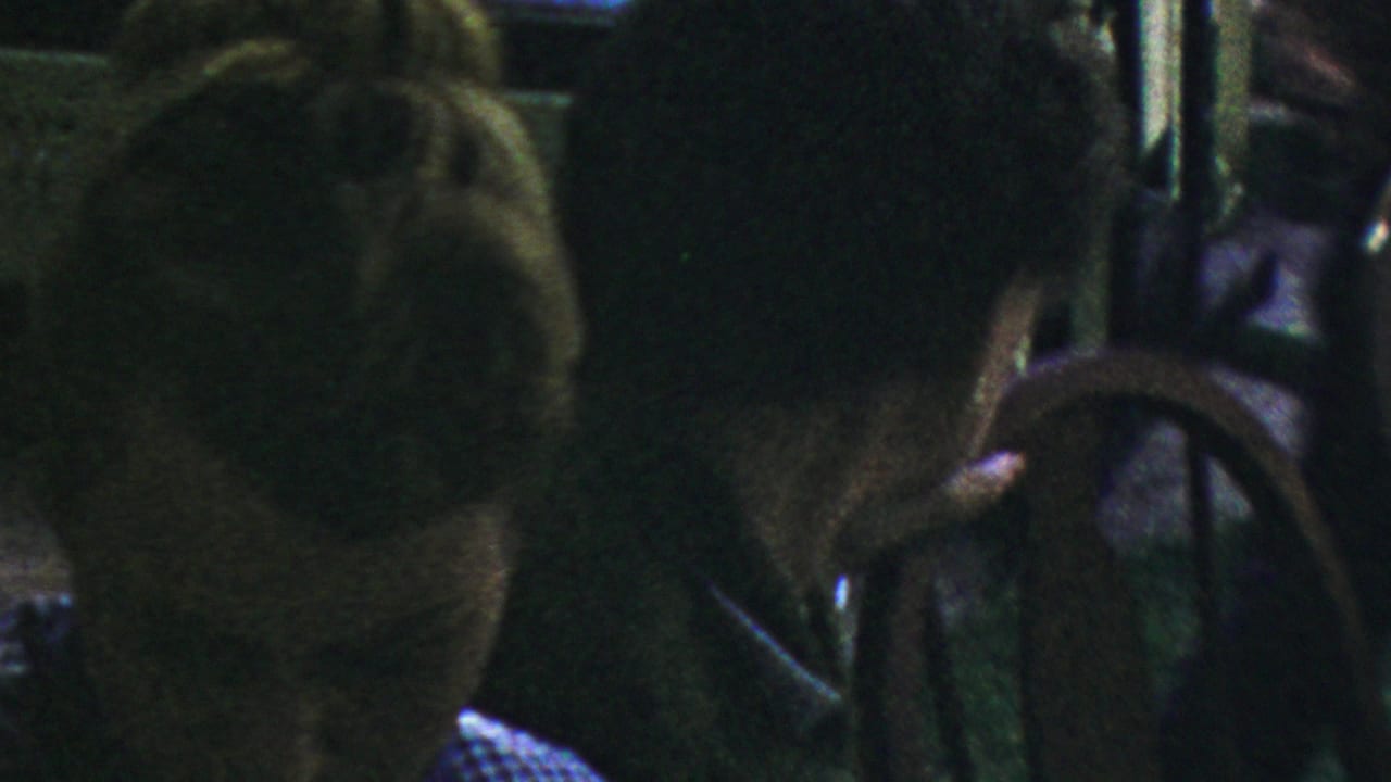 In a dark 1954 Columbus, Ohio restaurant home-movie frame, a woman with a mid-century updo sits near a man in a dark suit, with a chair back partly blocking the foreground.