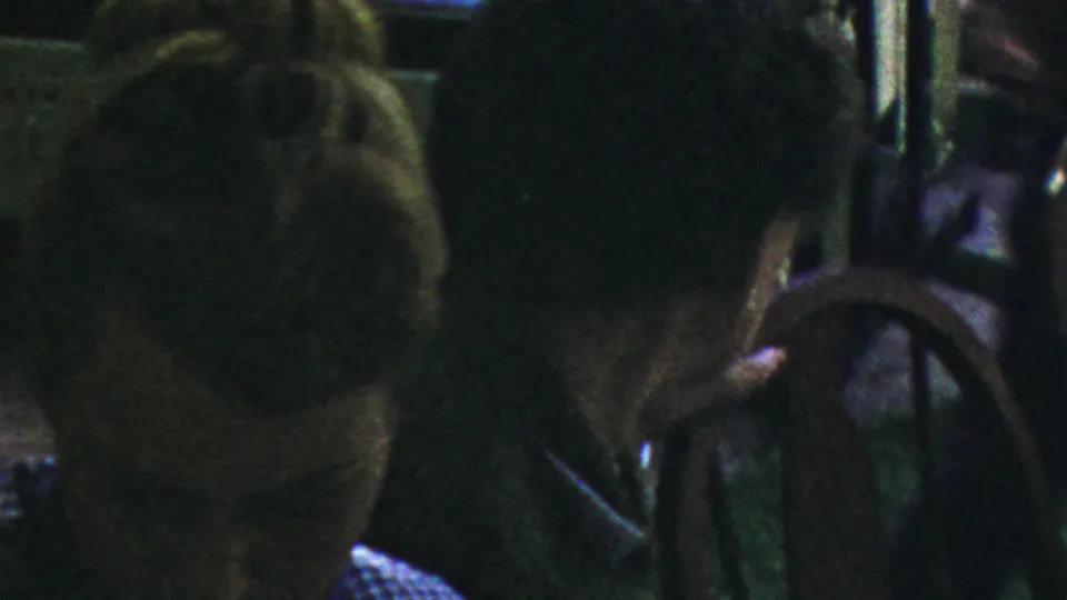In a dark 1954 Columbus, Ohio restaurant home-movie frame, a woman with a mid-century updo sits near a man in a dark suit, with a chair back partly blocking the foreground.