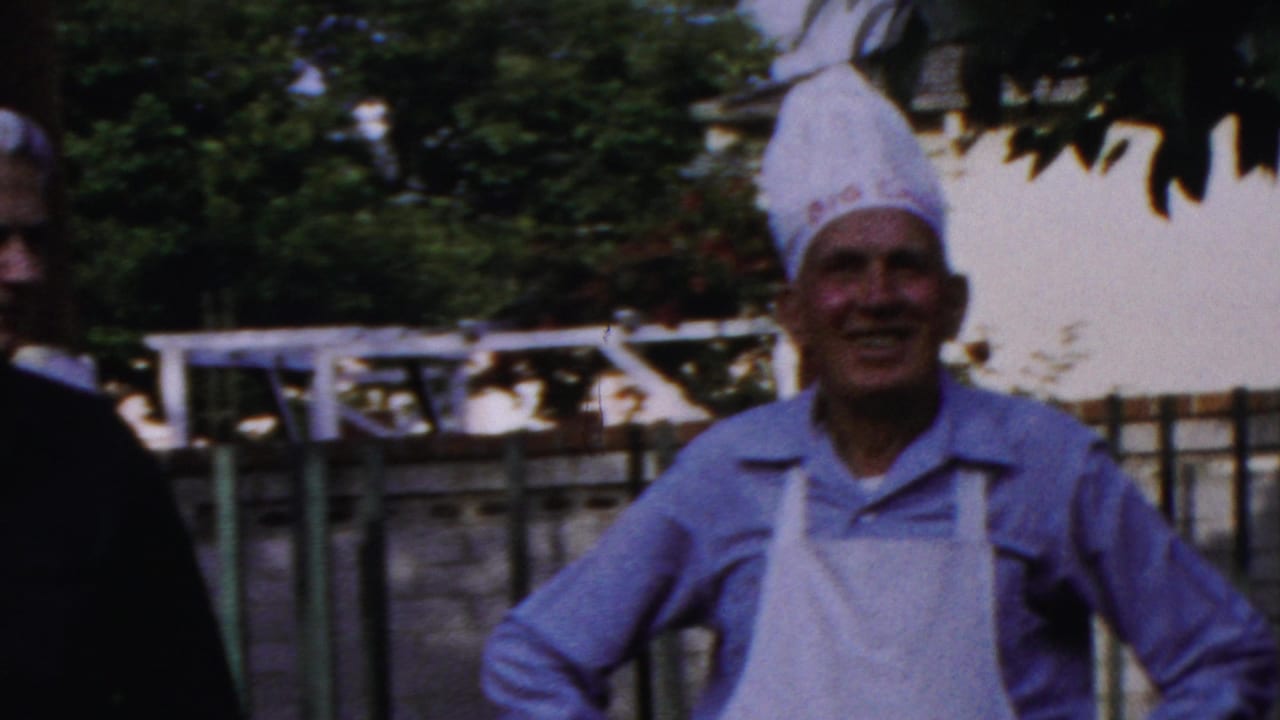 In Columbus, Ohio home-movie footage from 1954, adults stand in a backyard while a smiling man wears a white chef hat and apron near a woman at the edge of frame.