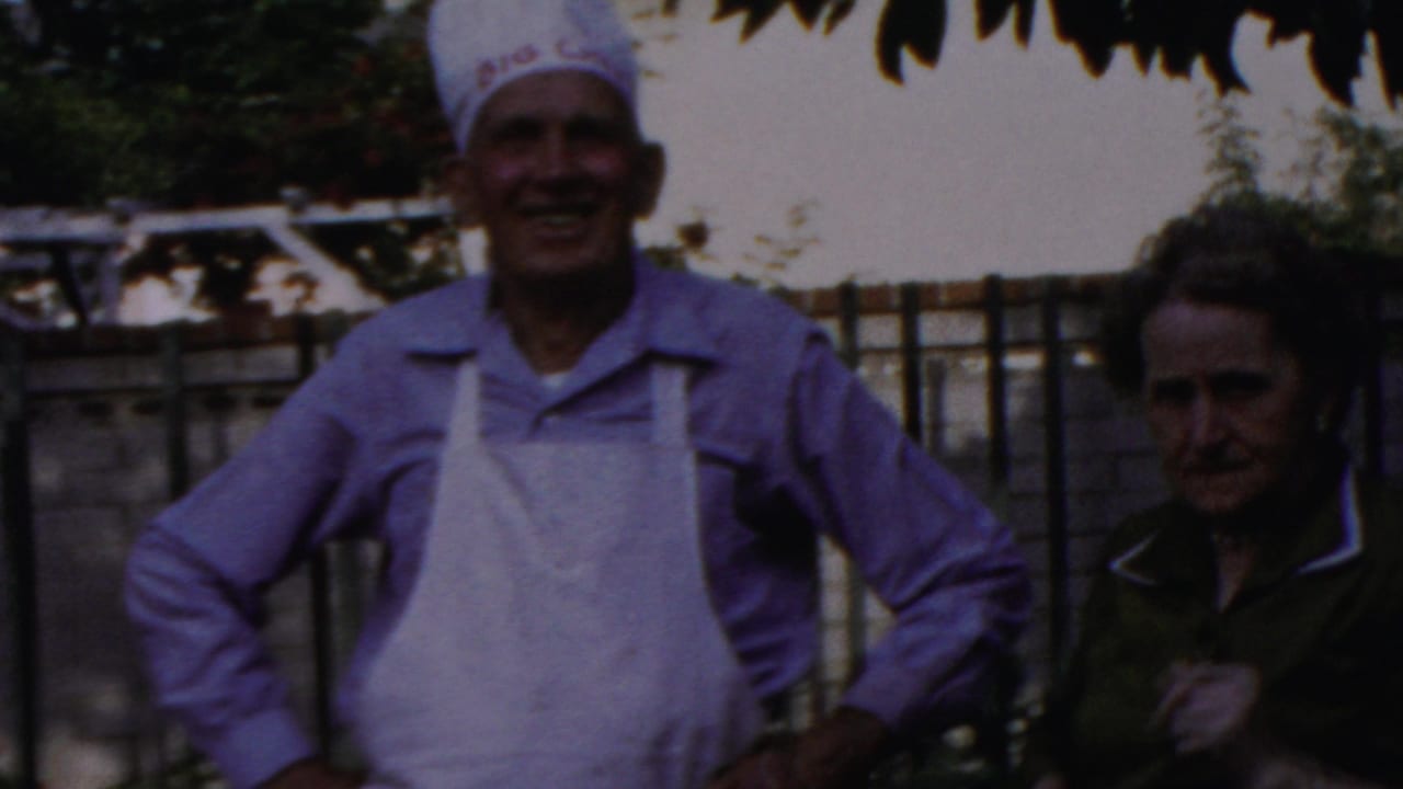 Smiling man in chef hat and apron stands with hands on hips beside a woman outdoors.