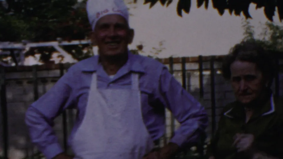 A smiling adult man in a white chef hat, purple shirt, and apron stands hands-on-hips in a Columbus backyard, with a woman visible beside him.