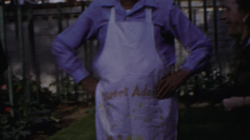 The man in chef hat and apron remains posed outdoors near a garden fence, with partial apron lettering visible but not clearly readable.