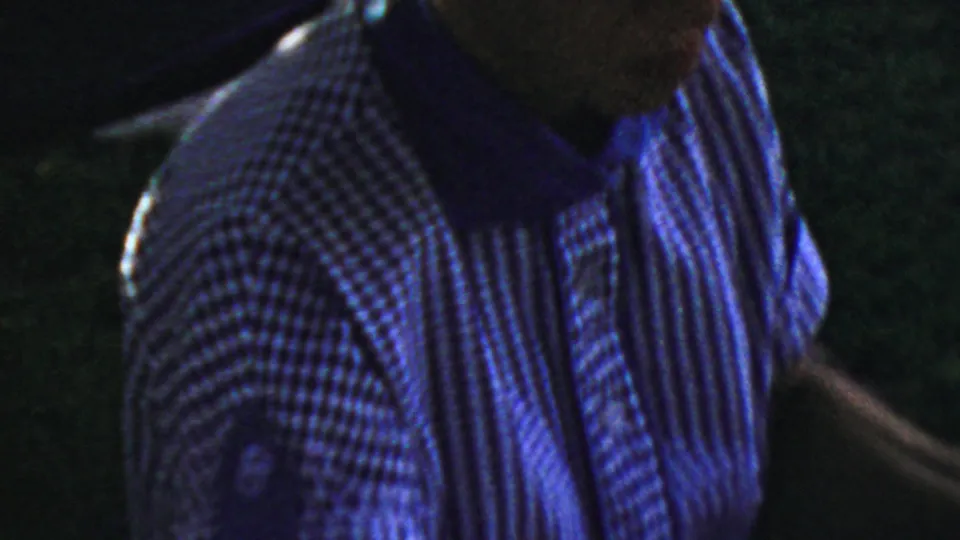 An adult in a striped shirt sits near the cluttered table in dim blue-tinted 1954 Columbus home-movie footage, with facial details partly obscured by low exposure.