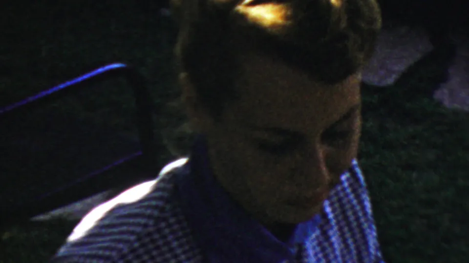 A woman in a striped shirt appears in profile beside the dark table during the 1954 Columbus cleanup scene, her face partially lit and softened by low-light 8mm grain.