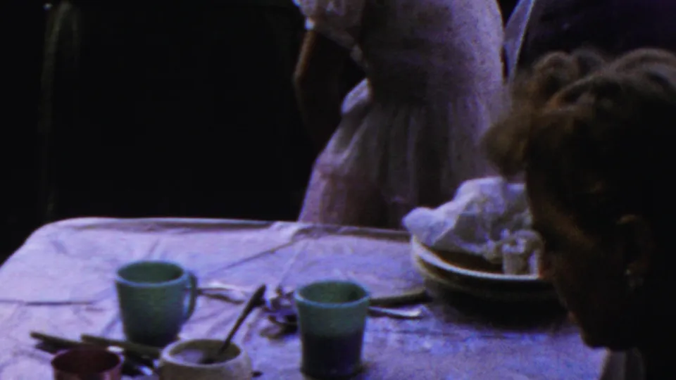 A table set with green cups, bowls, utensils, and plates is framed with partially visible people nearby in the Columbus, Ohio 1954 home movie.