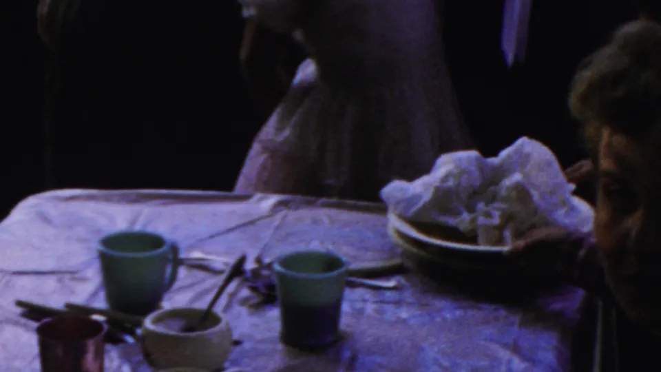 1954 Columbus, Ohio 8mm home-movie frame showing a dimly lit family dinner table with colored cups, plates, utensils, napkins, and dressed guests in the background.