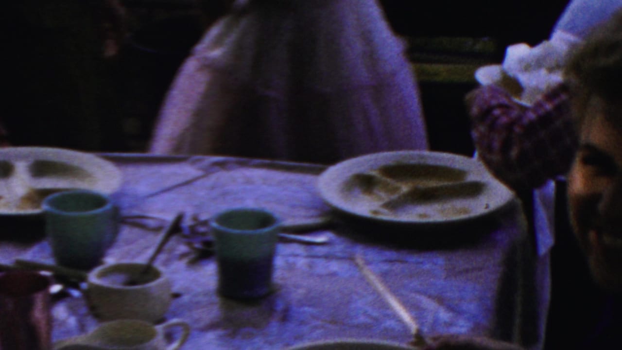 People gathered around a table with cups, plates, utensils, and a partially visible face near the center.