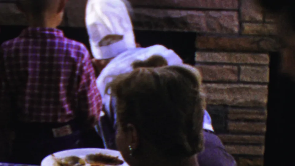 1954 Columbus, Ohio home-movie frame showing guests near a dinner table and brick fireplace area, with plates, cups, and several people partially visible from behind or in profile.