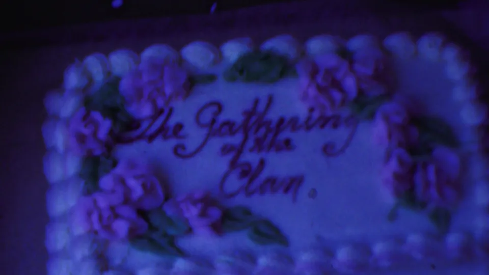 1954 Columbus, Ohio frame showing the decorated cake in soft focus, with purple flowers, green leaves, piped edging, and the phrase The Gathering of the Clan still visible.