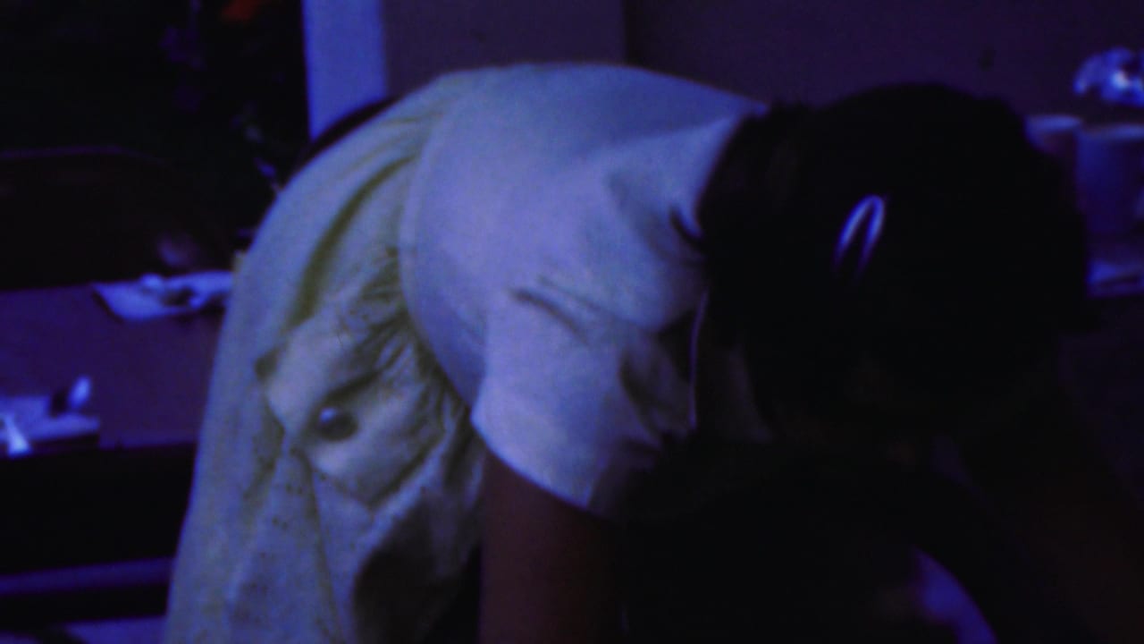 1954 Columbus, Ohio home-movie frame showing a woman in a light-colored dress bending forward inside a dim private room with household items behind her.