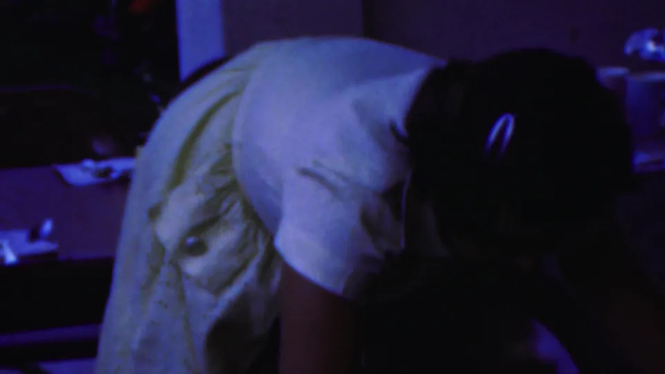 1954 Columbus, Ohio home-movie frame showing a woman in a light-colored dress bending forward inside a dim private room with household items behind her.