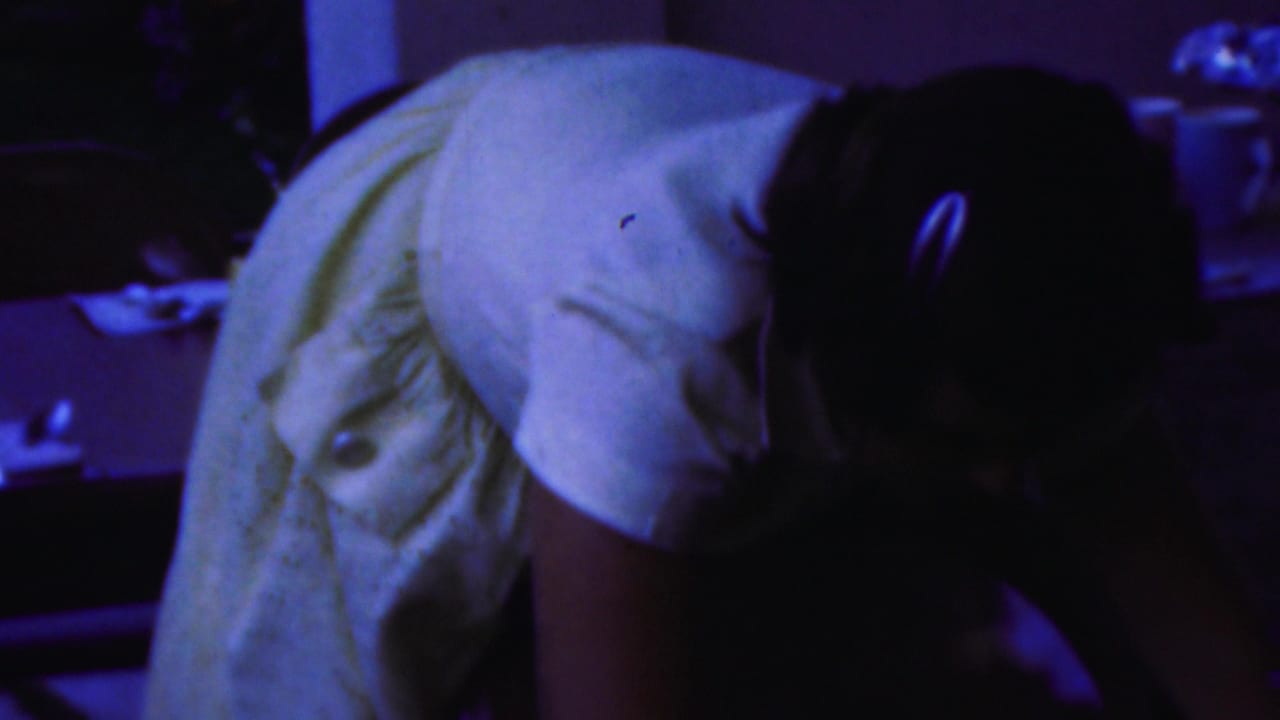 Woman in a light dress bending forward near a table in a dark room.
