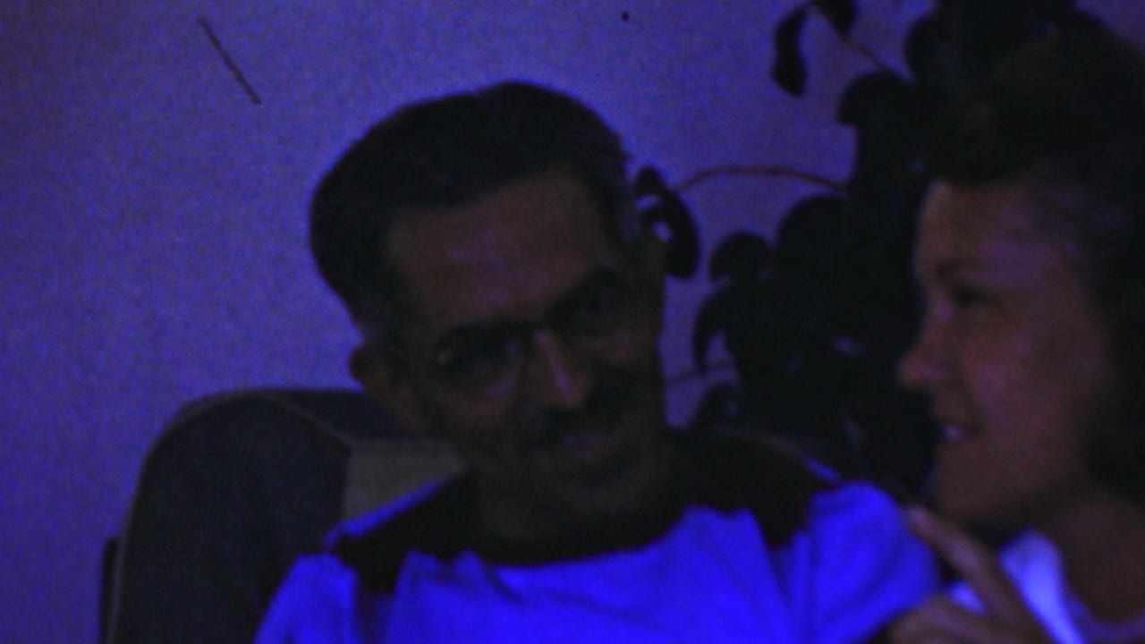 Man with glasses smiles beside a woman in a dim room.