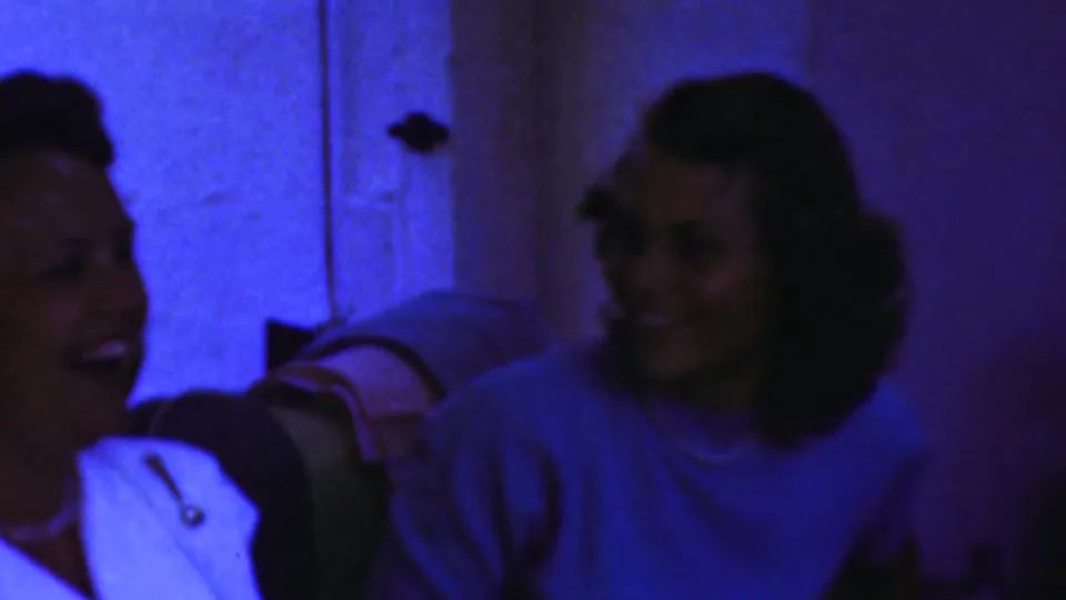 Two adult women laugh and face one another in a dim blue-lit private interior, part of 1954 Columbus, Ohio home-movie footage according to metadata.