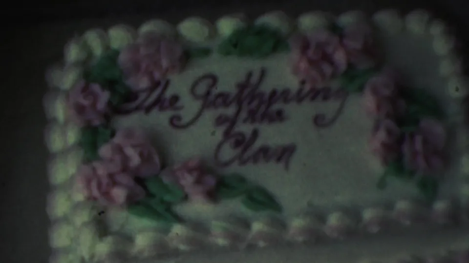 A close 8mm view shows the cake inscription The Gathering of the Clan, pink icing flowers, green leaves, and a white piped border, with no people visible in frame.
