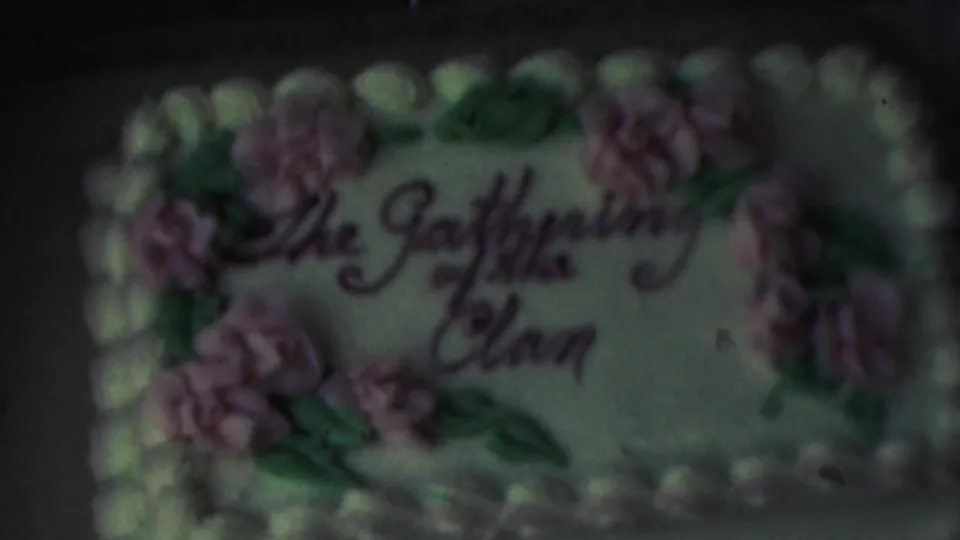 The final frame keeps the decorated cake centered, showing the clan-gathering inscription, floral frosting, muted color, and grainy 8mm home-movie texture.
