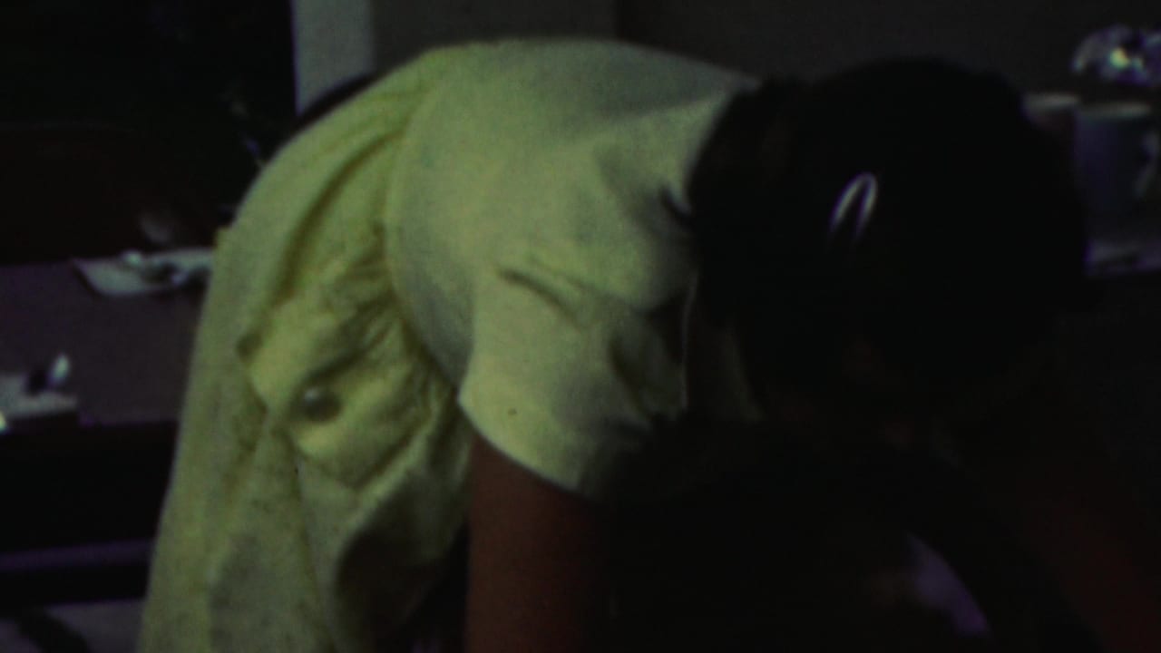 In 1949 Seaside, New Jersey home-movie footage, a woman in a pale dress bends forward near a table in a dim private interior.