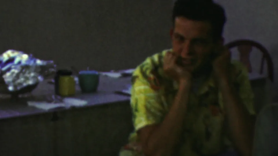A seated adult in a patterned yellow shirt smiles near a table with cups and foil-covered party items in the 1949 Seaside, New Jersey home movie.
