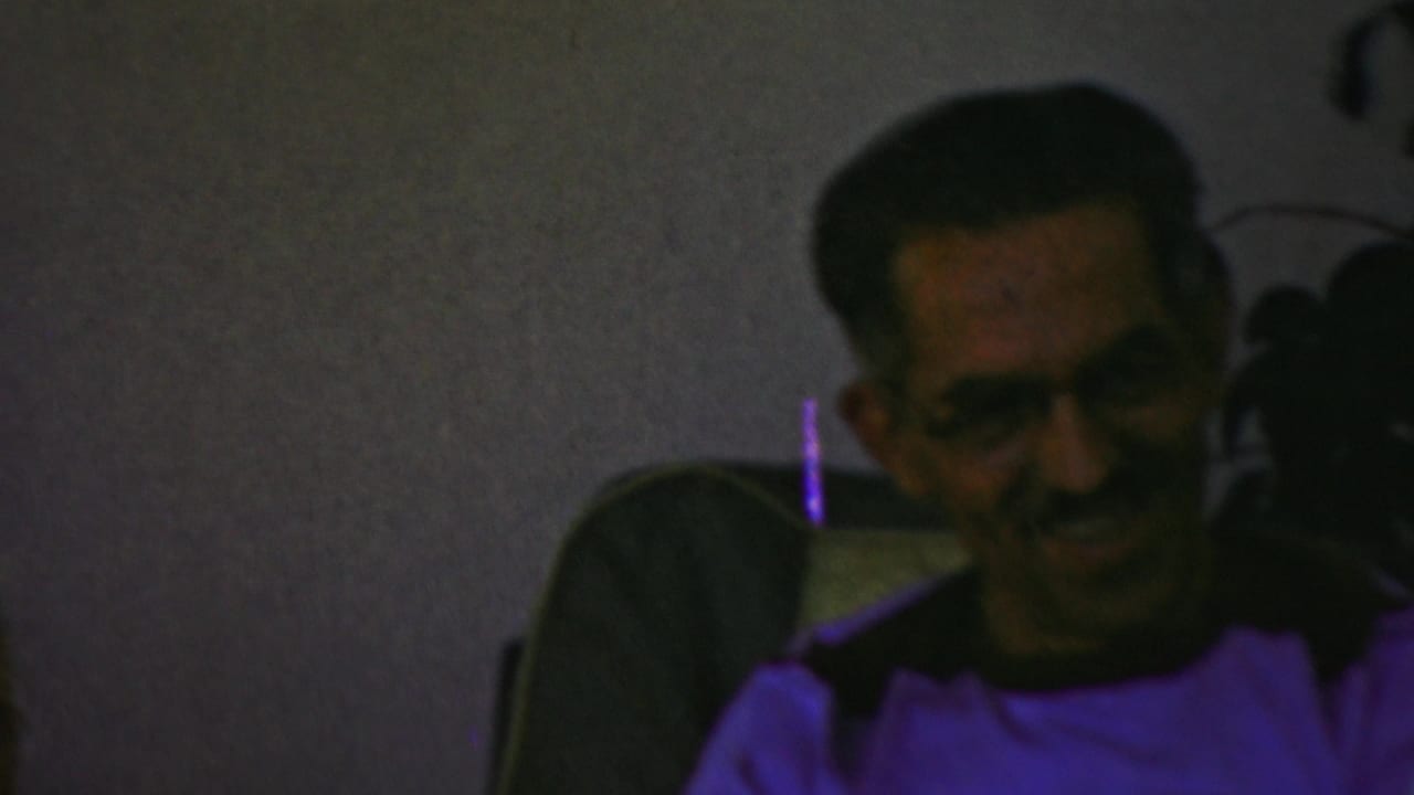 Man in glasses smiling while seated on a couch in a dim living room.