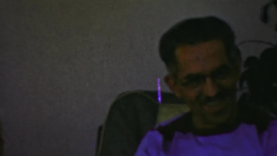 In the 1949 Seaside, New Jersey home-movie footage, a man wearing glasses sits on a couch and smiles in a dim private living-room interior.