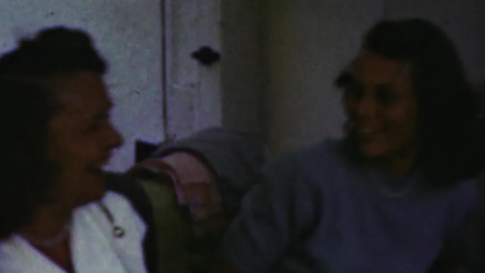 Two women are visible on a couch in a dim private living-room scene, with one smiling toward another person in the 1949 Seaside, New Jersey home-movie footage.