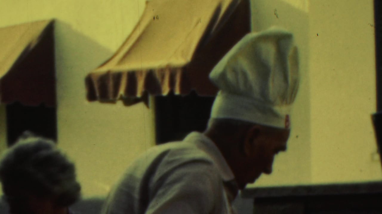 Man in a chef hat and apron seen beside striped awnings.