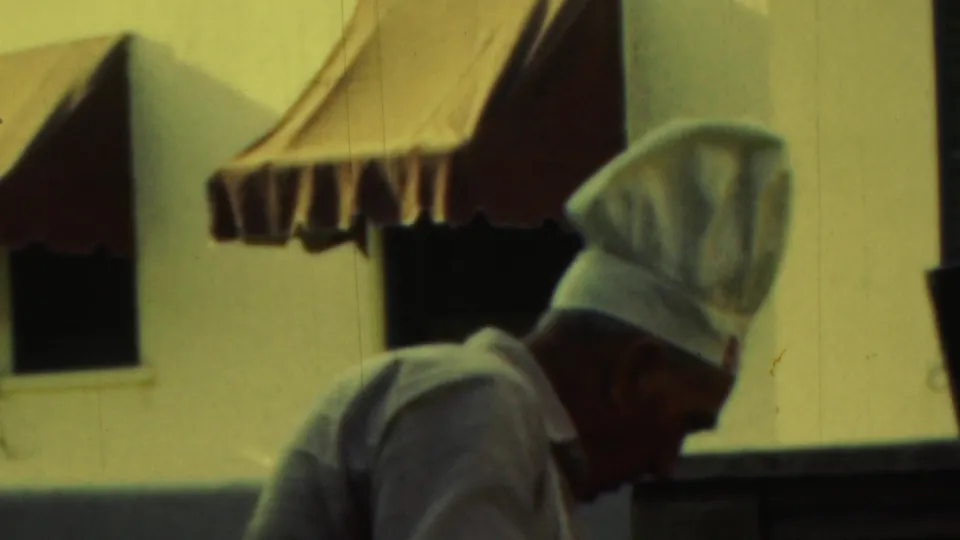 The chef-hatted man bends forward near a grill area or serving spot, with striped awnings and windows behind him in the 1949 home movie.