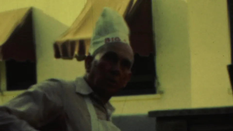 A man in a chef-style hat and apron turns toward the camera, with a light building wall and striped awnings behind him.
