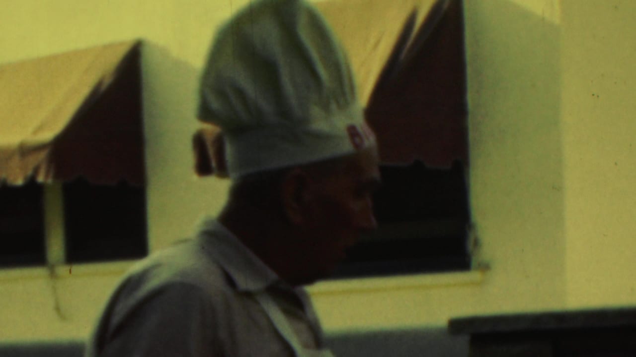 Man in a tall white chef hat stands beside a brick grill with awnings behind him.